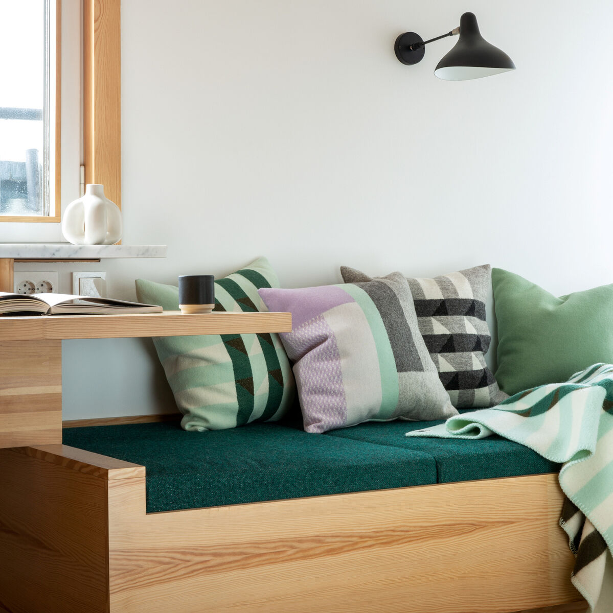 Ein gemütlicher Sitzbereich mit mehreren dekorativen Kissen in verschiedenen Farben und Mustern. Die Sitzbank und der Tisch bestehen aus hellen Holz und daneben befindet sich ein Fenster.