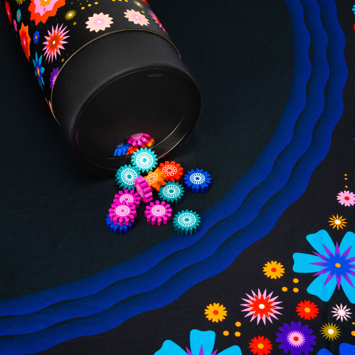 Schwarzes Spielfeld mit buntem Blumenmuster und einem offenen zylindrischen Behälter, aus dem farbige, gerillte Spielsteine auf das Spielfeld rollen. Die Szene zeigt ein modernes Brettspiel-Design mit kräftigen Farben und floralen Elementen entlang des Rands des Spielfeldes.