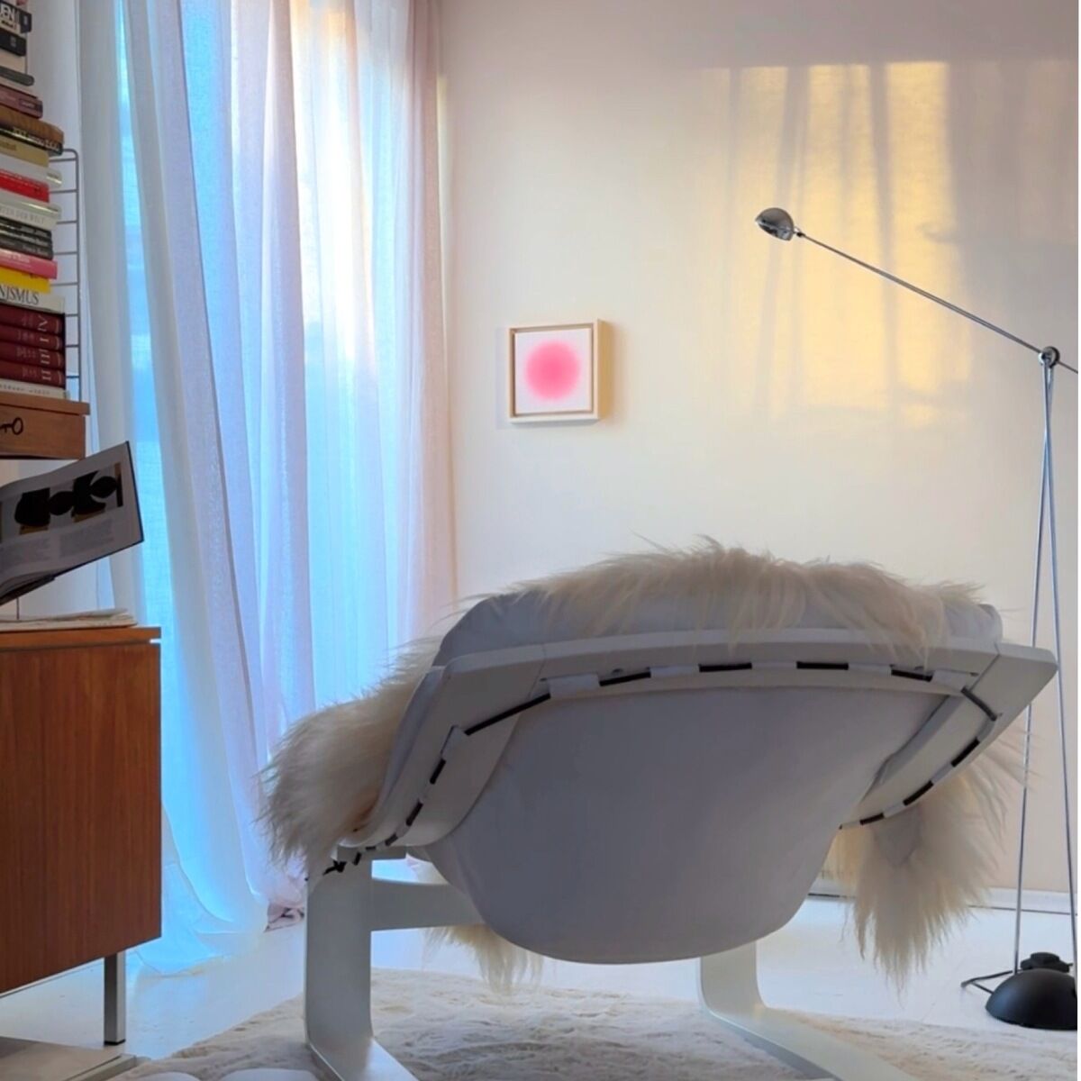Modern eingerichtetes Wohnzimmer mit weißem Designersessel, Schaffell, zahlreichen Büchern, Stehlampe und einem kleinen, quadratischen Kunstwerk mit rosa Farbverlauf (ZEIT-Sonderedition Sorbet »Pink«) an der Wand. Sanftes Tageslicht fällt durch halbtransparente Vorhänge und erzeugt eine ruhige, einladende Atmosphäre.