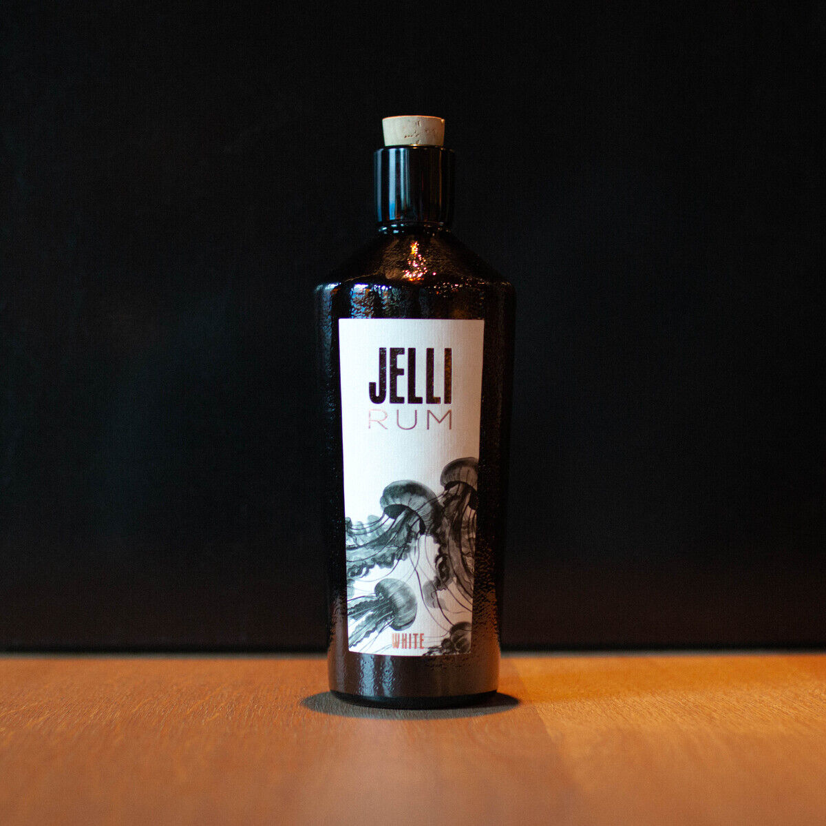 Die Abbildung zeigt eine Flasche »JELLI« Rum auf einer Holzplatte vor einem dunklen Hintergrund. Die Flasche ist dunkel mit einem weißen Etikett, auf dem der Name »JELLI« Rum zu sehen ist. Der Verschluss der Flasche ist aus hellem Holz.