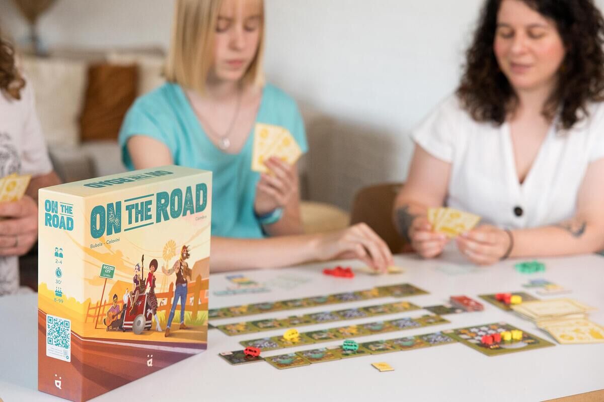 Drei Personen sitzen an einem Tisch und spielen das Brettspiel 'On the road'. Das Spiel beinhaltet Spielkarten und Spielstücke, die auf dem Tisch verteilt sind. Die Verpackung des Spiels steht ebenfalls auf dem Tisch.
