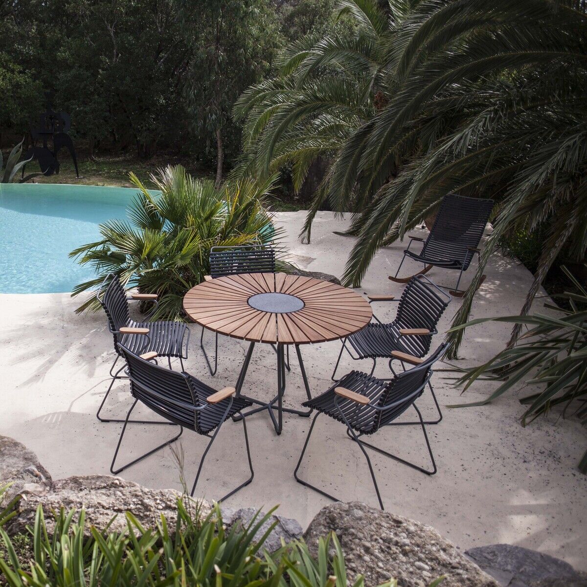 Ein Outdoor-Sitzbereich mit einem runden Holztisch und sechs CLICK Dining Chairs mit Armlehnen aus schwarzem Material, umgeben von üppigen grünen Pflanzen und einem einladenden blauen Pool im Hintergrund.