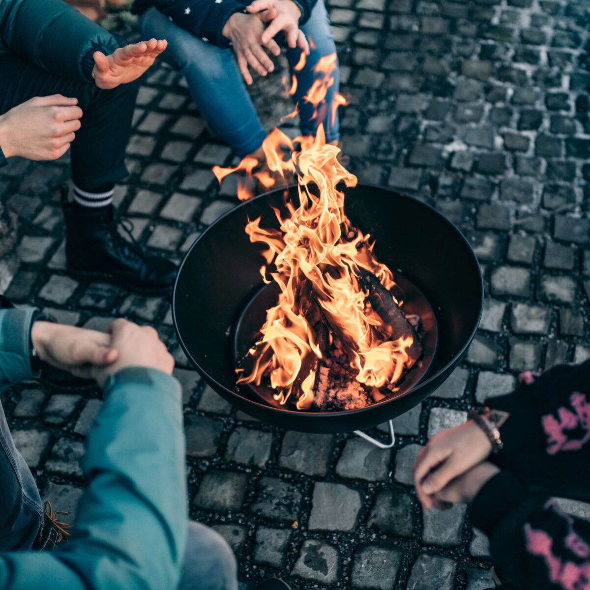Mehrere Personen sitzen um eine Feuerstelle im Freien; Flammen lodern auf Holzscheiten auf einem dunklen Bodenpflaster, entspannte Atmosphäre in einer Gruppenaktivität am Abend.
