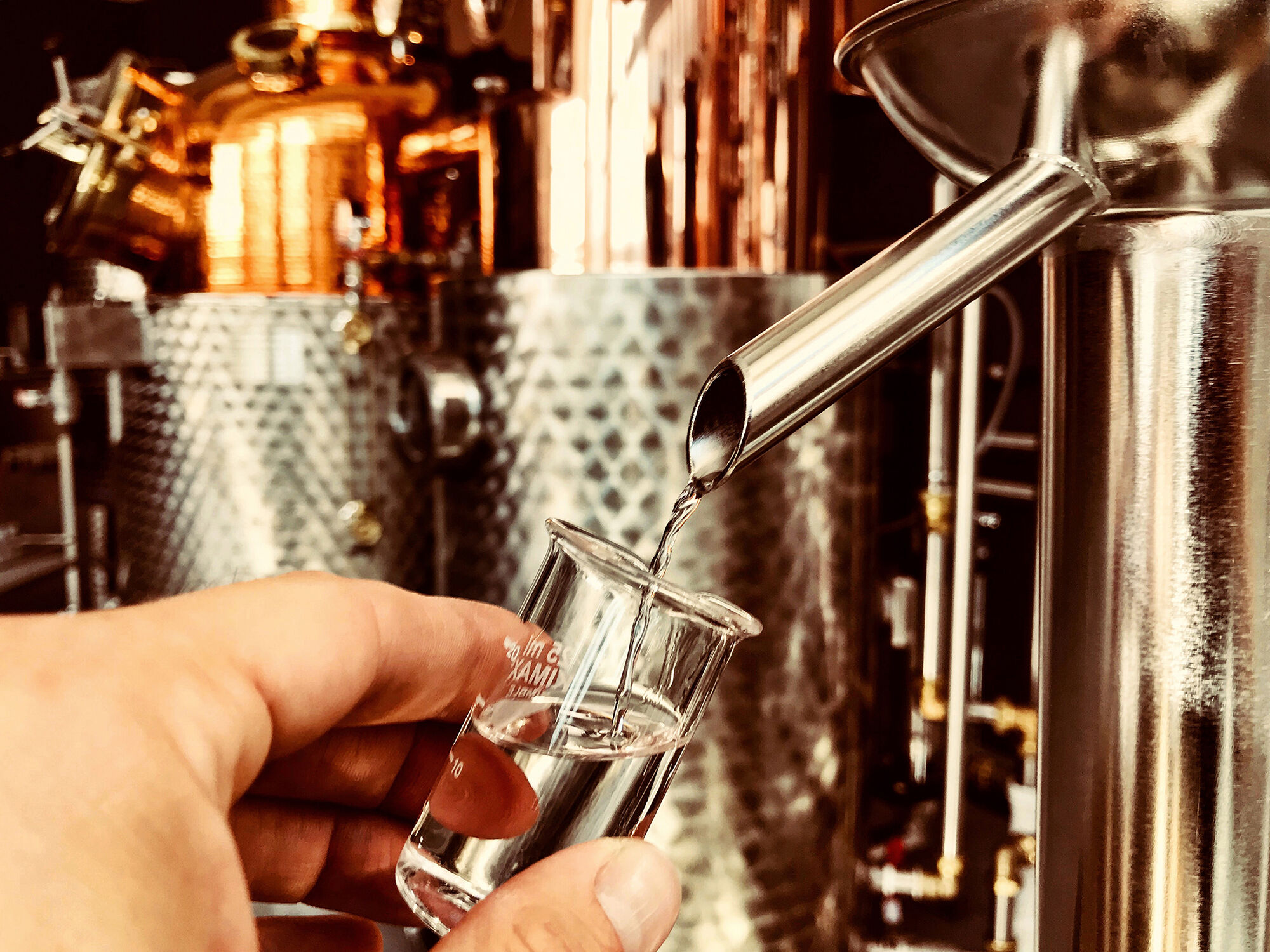 Hand hält ein kleines Glas, in das aus einer Metallrinne klare Flüssigkeit gegossen wird, vor einer Destillationsanlage