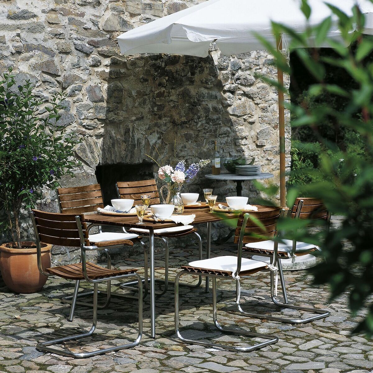 Gartentisch der Sonderedition von Thonet unter einem weißen Schirm im Außenbereich, umgeben von Stühlen und Pflanzen, auf einem Pflastersteinboden mit einer Steinmauer im Hintergrund.
