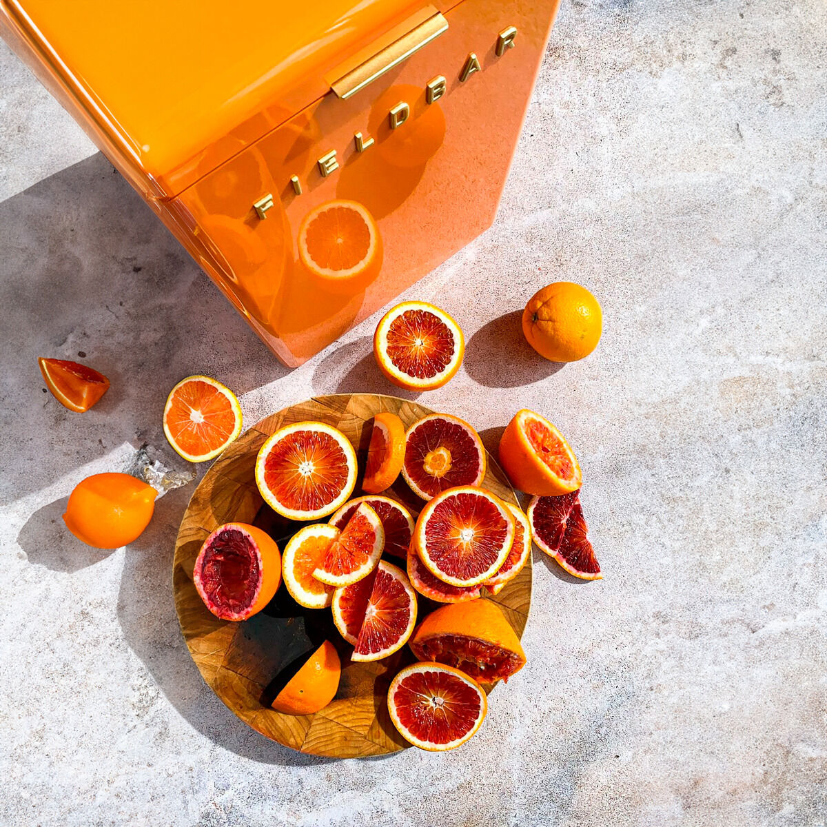 Eine orangefarbene Getränkebox neben einem runden Holzbrett mit aufgeschnittenen Orangen und Blutorangen. Einige ganze Orangen liegen verstreut auf dem hellgrauen Boden.