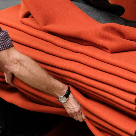 Ein Mann legt mehrere zusammengefaltete Merinodecken der Marke COASTLAND in glutroter Farbe mit anthrazitfarbenem Rand aufeinander. Im Hintergrund hängen weitere Decken. Die Szene zeigt hochwertige, weiche Wolltextilien im Herstellungsprozess.
