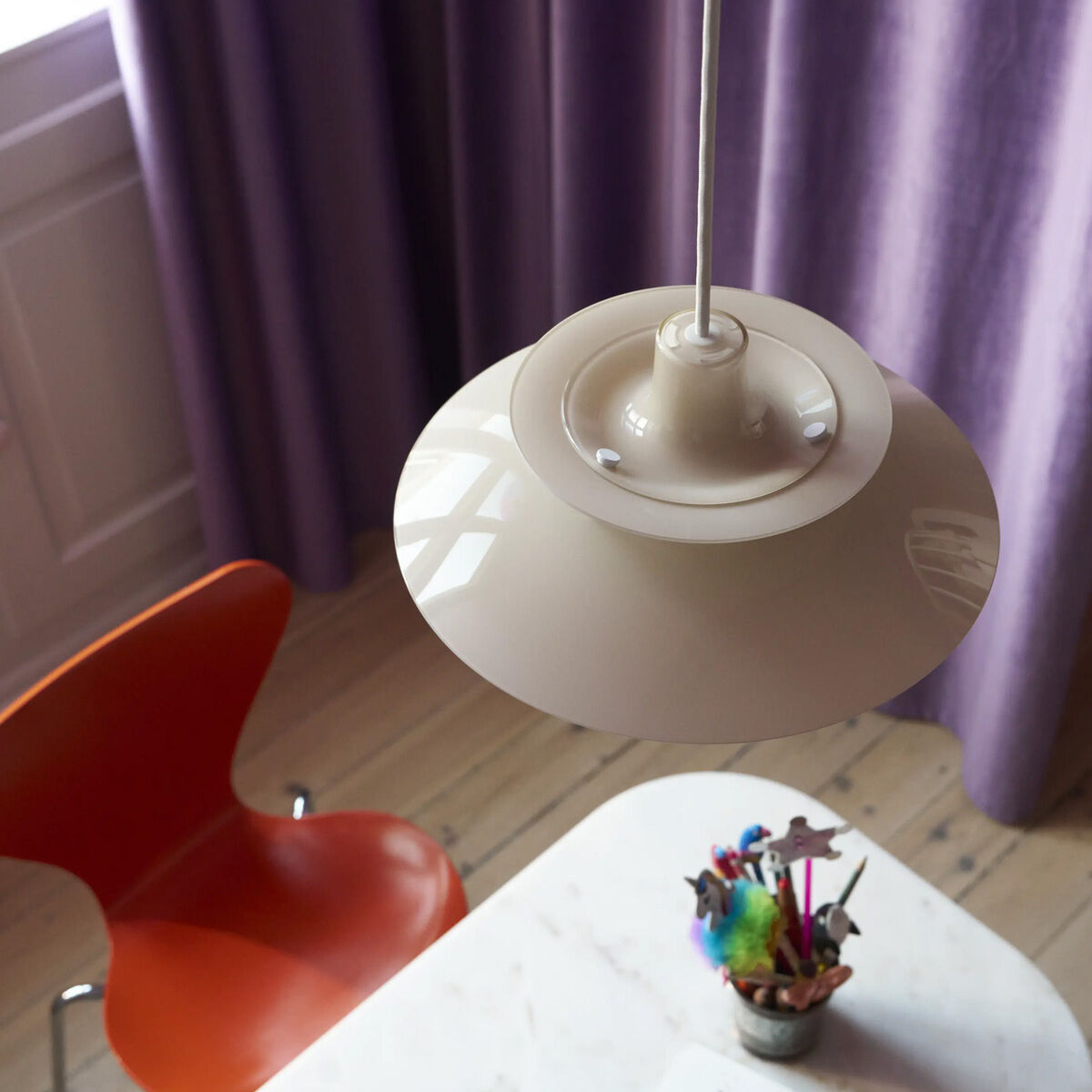 Die Hängeleuchte »PH 5 320« von Louis Poulsen in opal beige hängt über einem runden, weißen Tisch mit einer farbigen Vase. Links steht ein oranger Designstuhl, lila Vorhänge und helles Holzparkett sind im Hintergrund sichtbar.