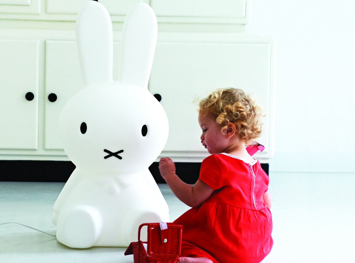 Ein kleines Kind sitzt auf einem weißen Boden und spielt mit einer roten Tasche. Daneben steht eine große weiße Lampe in Form des Cartoon-Charakters Miffy, die wie ein Hase aussieht.