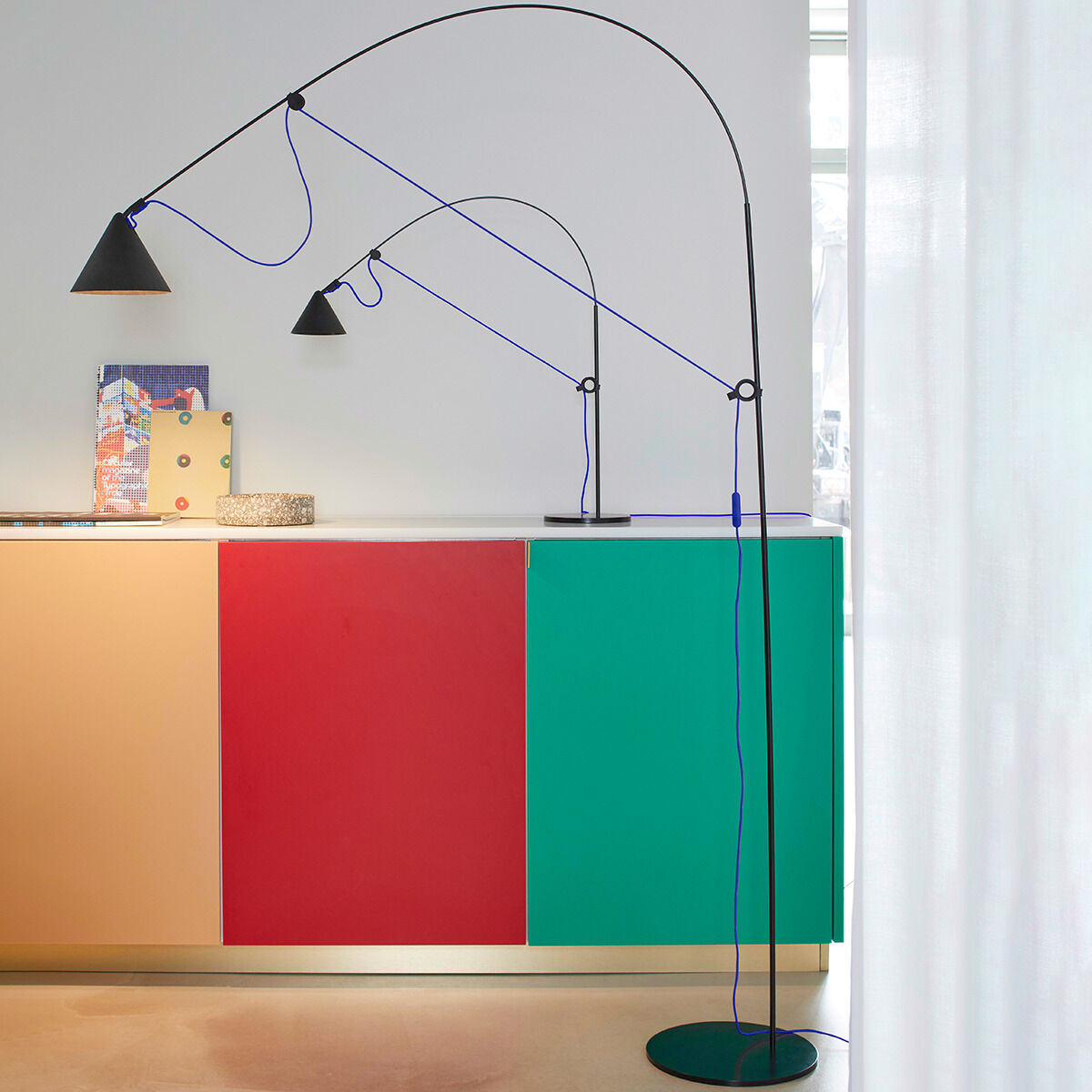 Eine moderne Stehlampe namens 'AYNO' in Schwarz/Ultramarin, platziert in einem stilvoll eingerichteten Raum. Die Lampe hat ein minimalistisches, elegantes Design mit einem verstellbaren Leuchtarm. Der Hintergrund zeigt farbige Paneele in Rot und Grün.