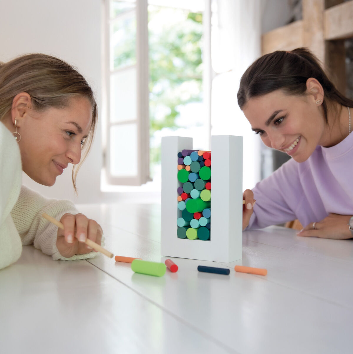 Zwei Frauen spielen ein buntes Geschicklichkeitsspiel namens 'Woodman' auf einem weißen Tisch. Das Spiel besteht aus einem weißen Rahmen, der mit bunten Holzstäbchen und -blöcken gefüllt ist.