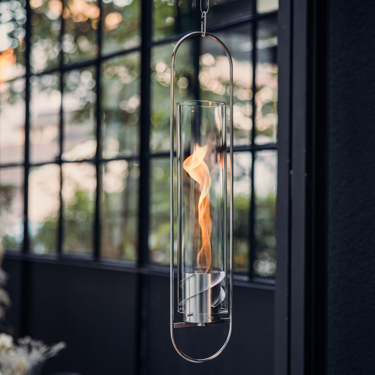 Das SPIN Hängesystem zeigt eine hängende Glas- und Metall-Laterne mit einer Flamme. Es ist in einem modernen Außenbereich aufgehängt und erzeugt eine warme, romantische Atmosphäre.