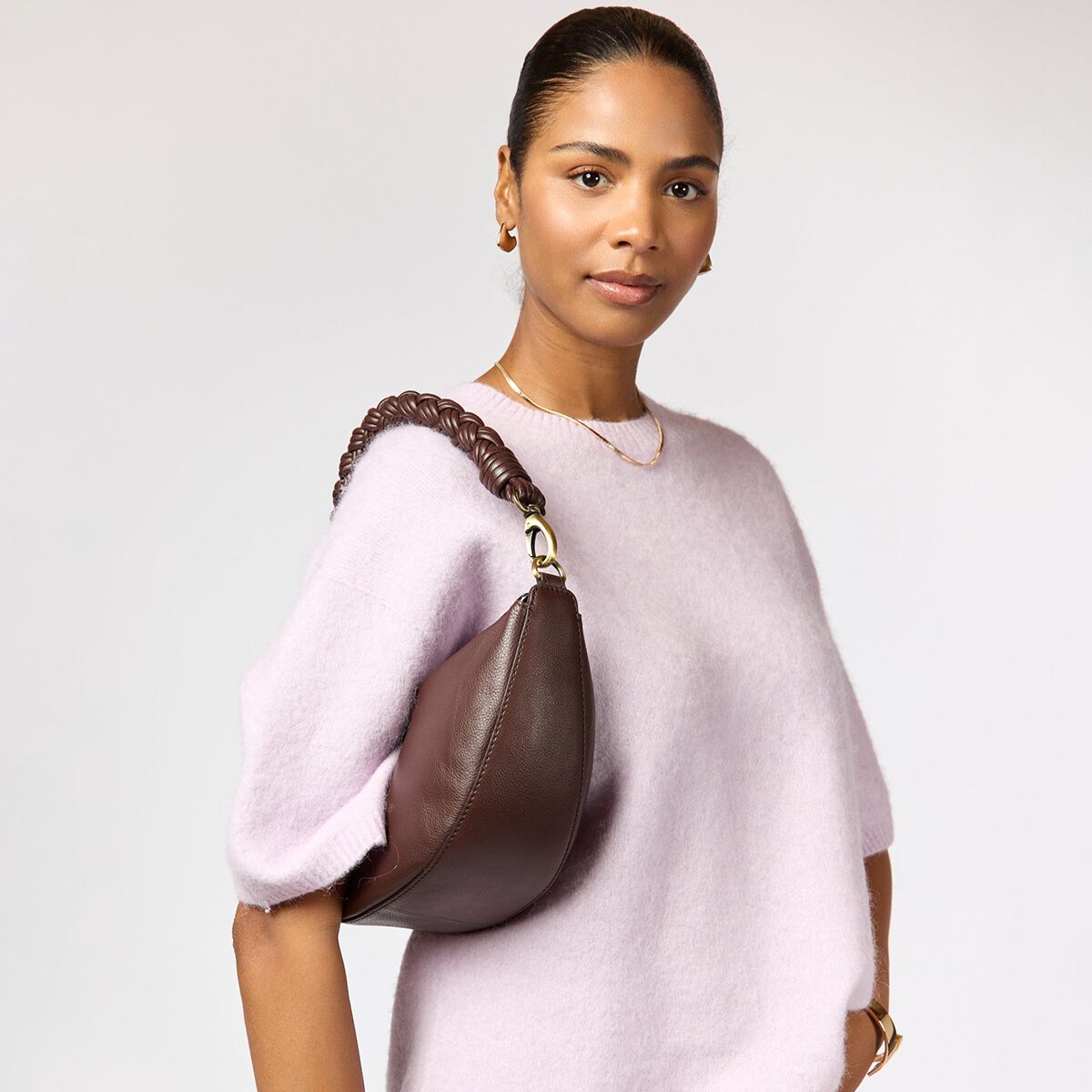 Eine Frau trägt die schokoladenbraune Crossbody-Tasche »Leo« von O My Bag aus offenporigem, umweltfreundlich gegerbtem Leder. Die Tasche hat einen geflochtenen Henkel und dezente Messingdetails. Sie wird über einem weiten, hellvioletten Pullover getragen und wirkt modern und elegant.