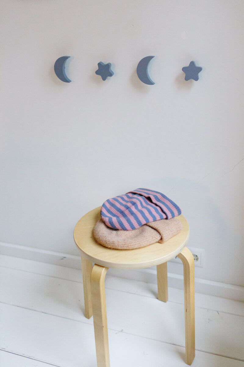 Drei an der Wand montierte Haken in Form von blauen Sternen und Monden über einem kleinen Holzhocker mit rosa und blau gestreiften Kleidungsstücken auf einem beigen Tuch.