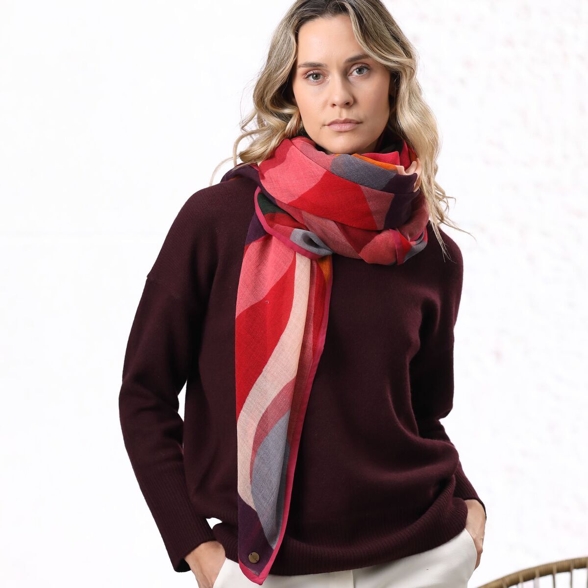 Eine Frau trägt einen mehrfarbigen Wollschal namens 'Yara', der in verschiedenen Rot- und Grautönen gestaltet ist. Sie kombiniert den Schal mit einem dunklen Pullover und einer hellen Hose. Der Schal liegt locker und stilvoll um ihren Hals.