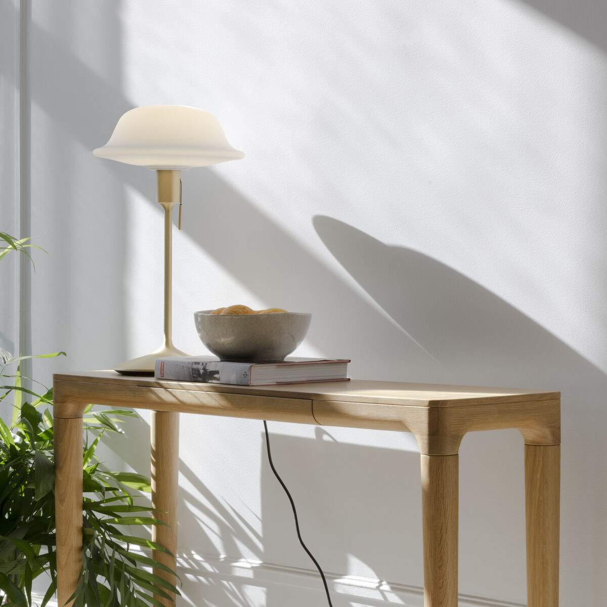 Eine helle, minimalistisch eingerichtete Ecke mit der Konsole »Heart'n'Soul« von Umage aus hellem Holz. Darauf stehen eine weiße Design-Tischleuchte, ein Buch, eine dekorative Schale und eine große grüne Zimmerpflanze. Sonnenlicht erzeugt Schatten an der weißen Wand.
