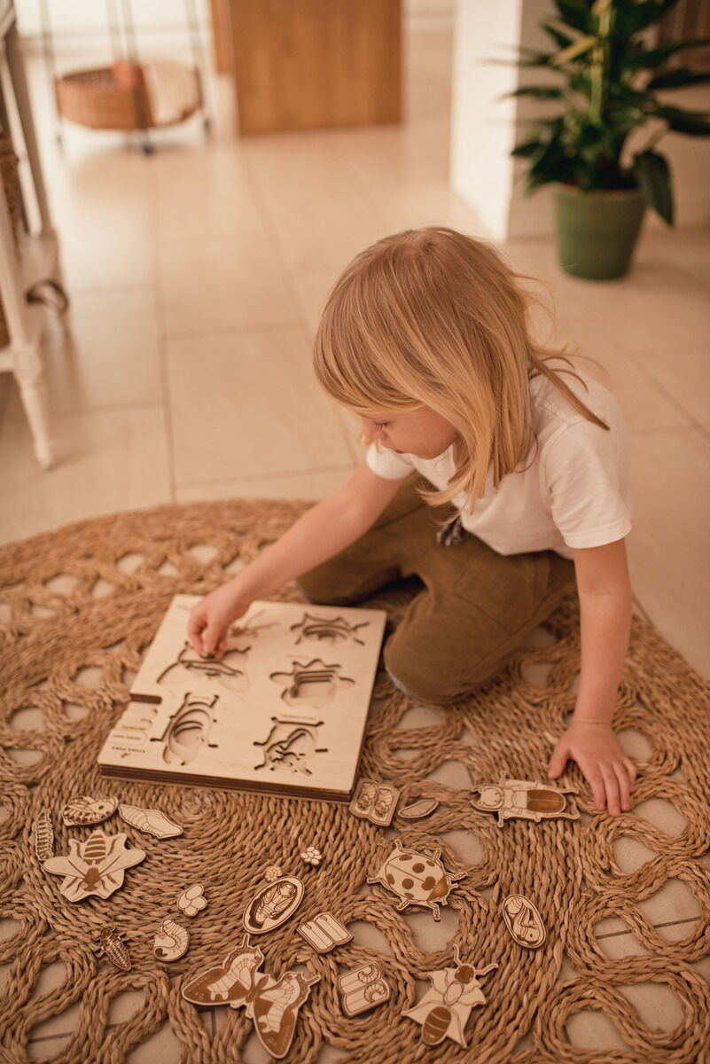 Ein Kind spielt auf einem Teppich mit dem Holzpuzzle »Leben des Käfers«. Das Puzzle zeigt verschiedene Käfer und ist aus naturfarbenem Holz gefertigt.