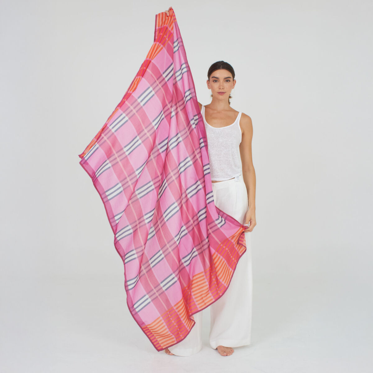 Frau präsentiert den großen Baumwoll-Seidenschal »Morgan« von Shalls Barcelona. Das Tuch zeigt ein auffälliges Karomuster in Rosa, Pink, Orange und Rot. Die Frau trägt weiße, fließende Kleidung. Der Hintergrund ist neutral weiß gehalten und hebt das bunte Muster hervor.