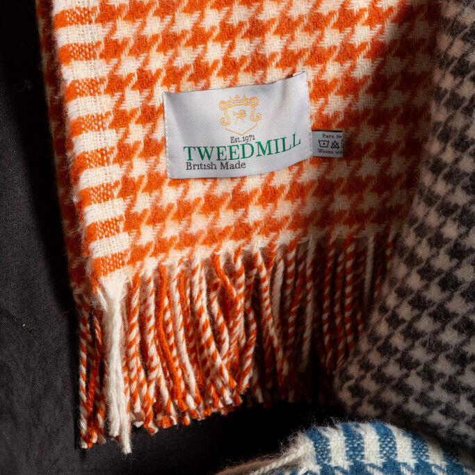 Eine Decke mit Hahnentritt-Muster, die vor allem in Orange und Weiß gestaltet ist. Sie hat Fransen an den Enden und ein Etikett, das auf britische Herstellung hinweist.