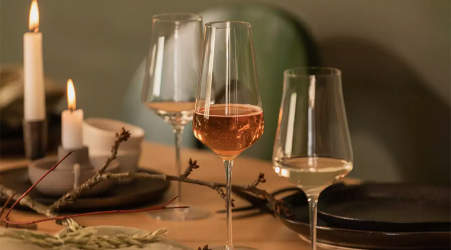 Drei Weingläser mit rosé und weißem Wein auf einem gedeckten Tisch mit brennenden Kerzen und dekorativen Zweigen