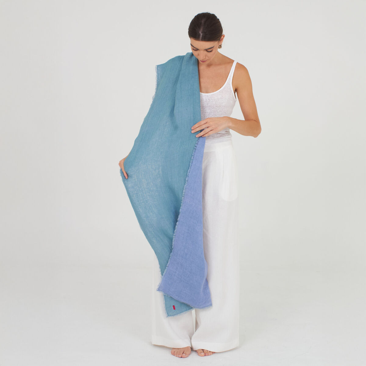 Frau steht barfuß vor weißem Hintergrund und trägt einen langen, zweifarbigen Leinenschal »Alsi« von Shalls Barcelona in oceantönen. Der Schal liegt locker über ihrer Schulter und reicht fast bis zum Boden. Sie trägt ein weißes Top und weite weiße Hosen.