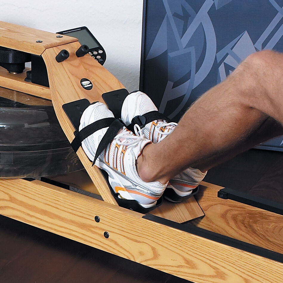 Eine Person mit Sportschuhen sitzt auf einem Waterrower-Rudergerät aus Eichenholz. Ein schwarzes Bedienfeld ist oben am Gerät befestigt. Im Hintergrund sind dunkle grafischen Muster an einer Wand zu sehen.