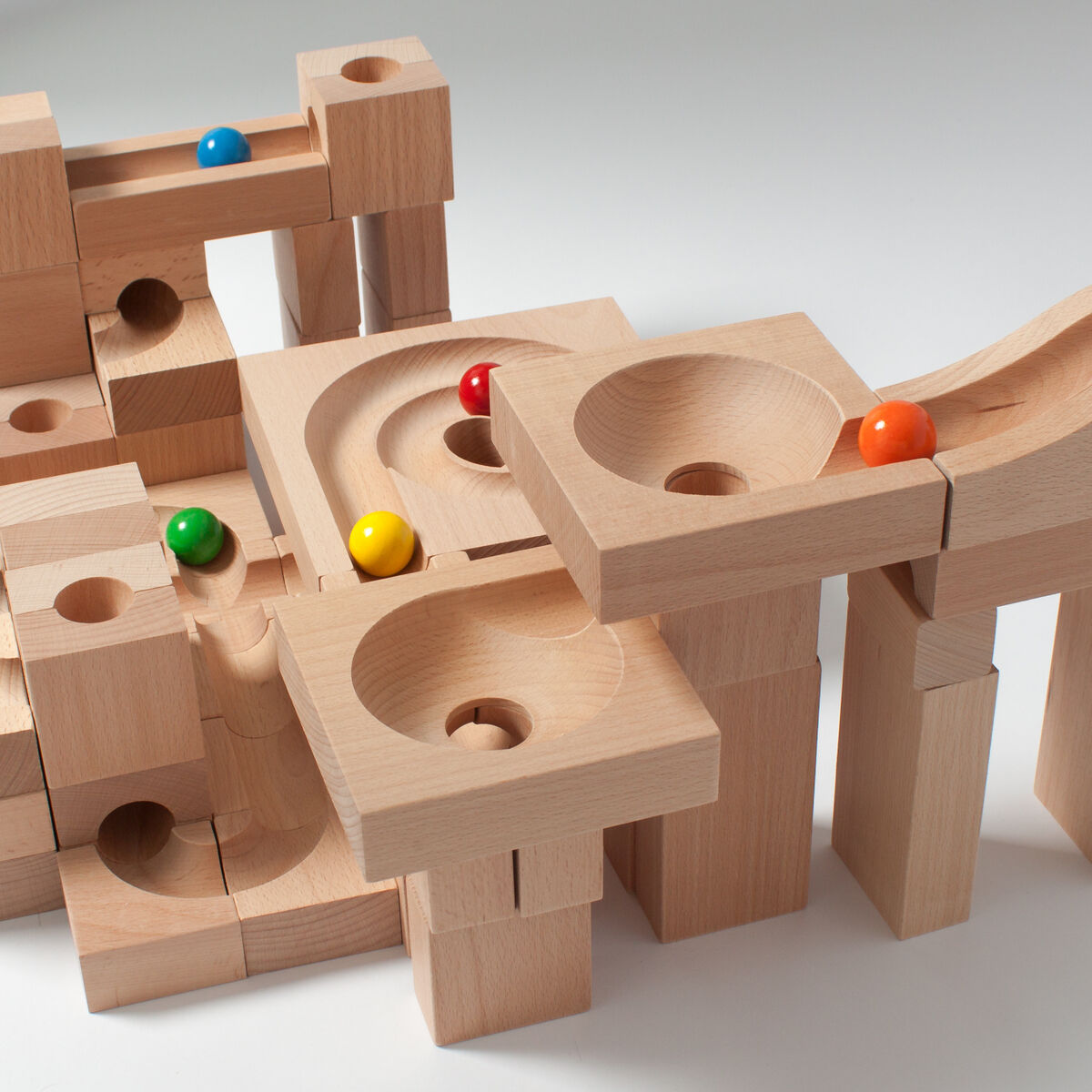 Eine Kugelbahn aus Holz mit bunten Kugeln. Die Konstruktion besteht aus mehreren hölzernen Bauklötzen, die so angeordnet sind, dass die Kugeln durch die Bahn rollen können. Die Kugeln haben die Farben Rot, Blau, Grün, Gelb und Orange.
