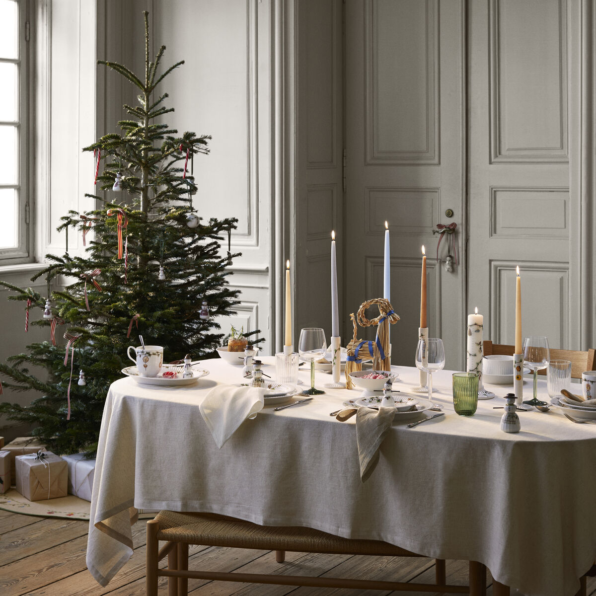 Ein festlich gedeckter Tisch in einem elegant eingerichteten Raum mit einem geschmückten Weihnachtsbaum im Hintergrund. Auf dem Tisch und am Tannenbaum befindet sich unter anderem ein kunstvoll gearbeiteter Anhänger in Form eines Schneemädchens als winterliche Dekoration.
