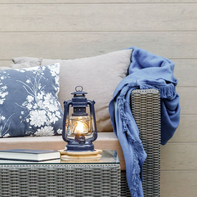 Eine LED-Laterne namens 'Baby Special 276' steht auf einer geflochtenen Outdoor-Couch mit grauen Kissen und einer blauen Decke. Im Hintergrund eine Holzverkleidung.
