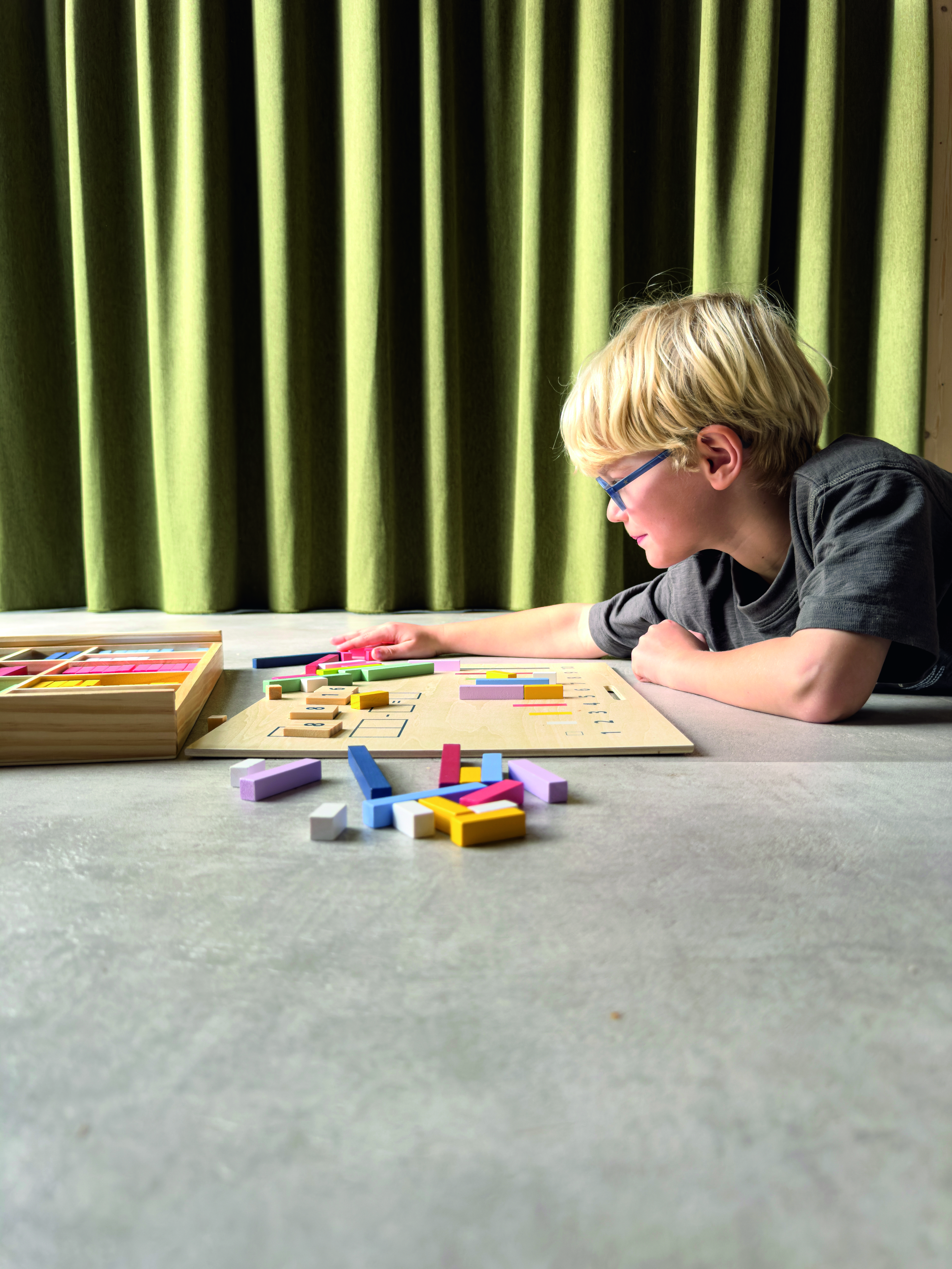 Ein Kind spielt am Boden mit einer Lernbox, die Rechenstäbchen und Zahlen enthält. Das Set besteht aus verschiedenfarbigen Holzstäbchen, einer Holzplatte und einer Holzbox. Die Lernumgebung ist hell und gemütlich mit einem Vorhang im Hintergrund. Das Produkt fördert das mathematische Verständnis und mathematische Fähigkeiten, besonders für Vorschulkinder und Grundschüler.