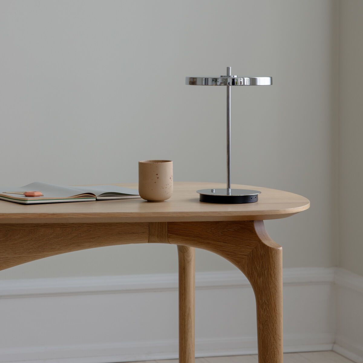 Auf einem hellen Holztisch steht eine moderne, minimalistische Tischlampe mit poliertem Stahlfuß. Daneben befinden sich eine beige Tasse und ein offenes Buch. Die Wand im Hintergrund ist hell, das Gesamtbild wirkt ruhig und zeitlos.