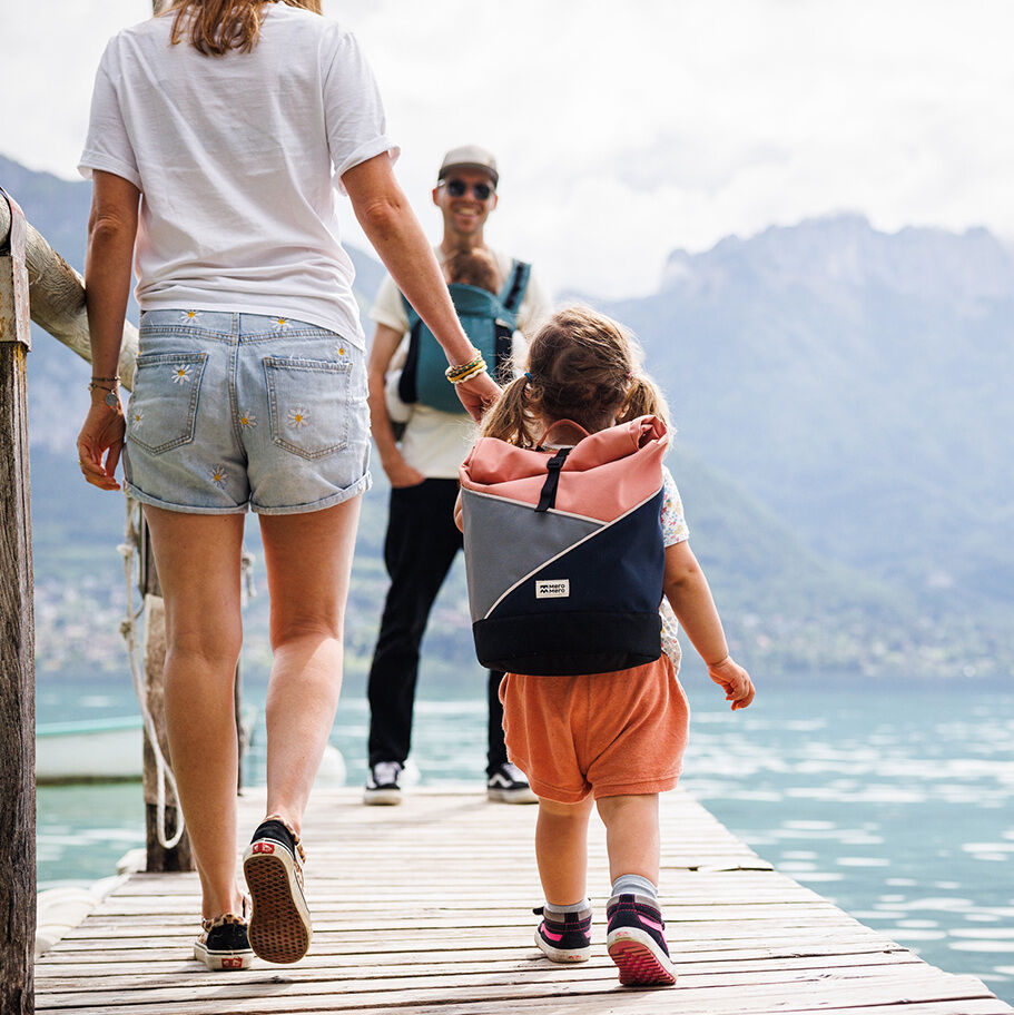 Ein Kind trägt einen Rucksack der Marke Popoyo und spaziert auf einem Holzsteg am See. Zwei Erwachsene begleiten das Kind.