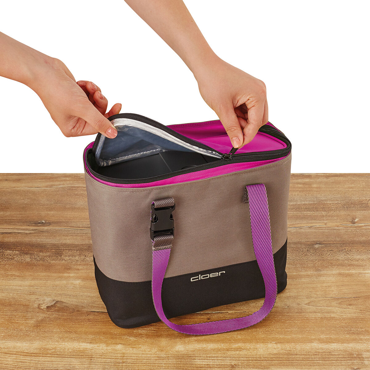Eine Lunch-Tasche mit grauer Außenseite, schwarzem Boden und pinkfarbenen Griffen und Reißverschluss. Zwei Hände öffnen die Tasche, die auf einer Holzoberfläche steht.