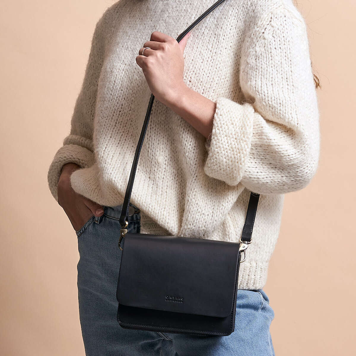 Eine Frau trägt eine cremefarbene Strickjacke und eine hellblaue Jeans, dazu eine kleine schwarze Leder-Schultertasche namens 'Audrey Mini'.