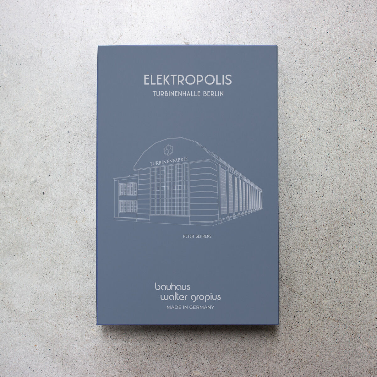 Die Verpackung der ZEIT-Sonderedition Uhr »Elektropolis« Oliv ist in elegantem Grau gehalten, zeigt eine skizzierte Turbinenhalle Berlin und weiße Schrift. Oben stehen Titel und Untertitel, unten das Bauhaus-Logo, Hersteller und 'Made in Germany'.