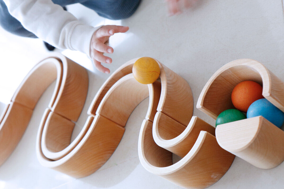 Ein Kind spielt mit einem biegsamen Holzspielzeug. Das Spielzeug besteht aus natürlich gefärbtem Holz und umfasst verschiedene farbige Kugeln in Gelb, Grün und Blau, die durch einen Holzweg rollen.