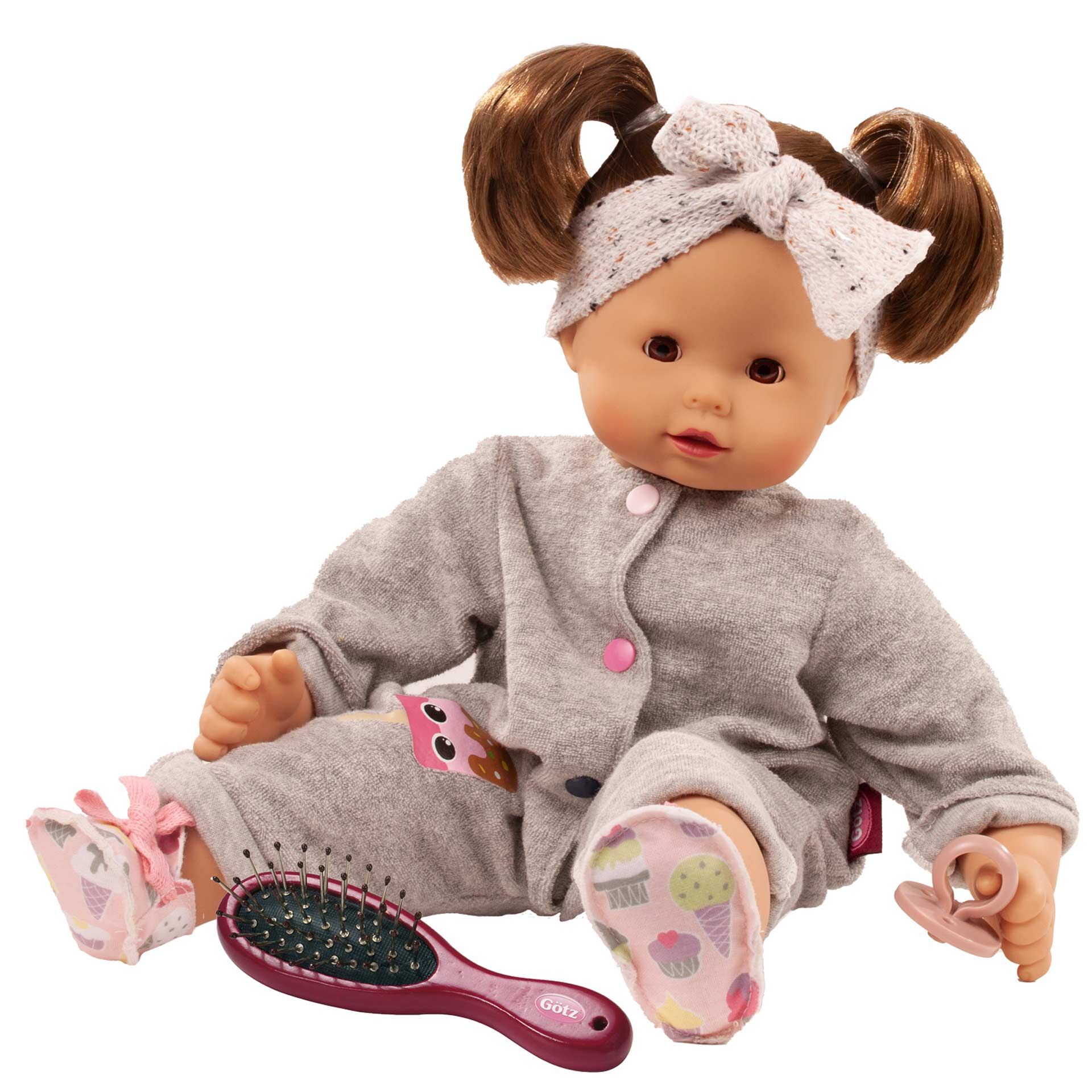 Weiche Babypuppe mit braunen Zöpfen, grauem Schlafanzug und rosa Haarschleife, sitzt aufrecht, trägt bunte Söckchen und hält eine kleine Haarbürste. Kindgerechtes Design und liebevolle Details, ideal als Kuschelpuppe oder Spielzeug für Kleinkinder.