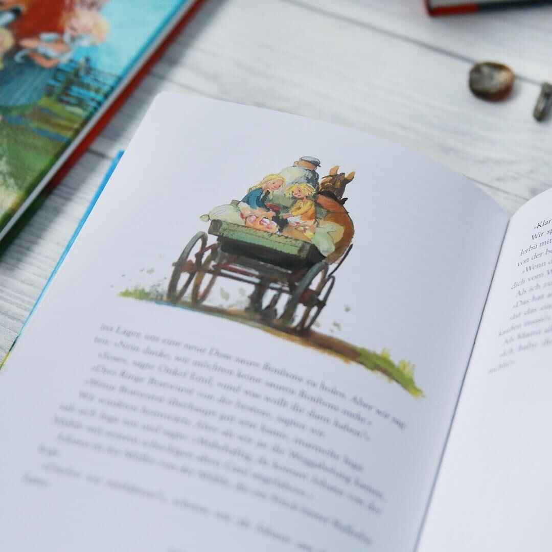 Ein offen liegendes Buch mit Illustrationen aus dem Kinderbuch 'Wir Kinder aus Bullerbü' von Astrid Lindgren. Die gezeigte Illustration zeigt Kinder in einem Wagen, die von einem Erwachsenen gezogen werden. Im Hintergrund ist ein zweites Buch zu sehen sowie ein heller Holztisch und Dekorationsgegenstände.