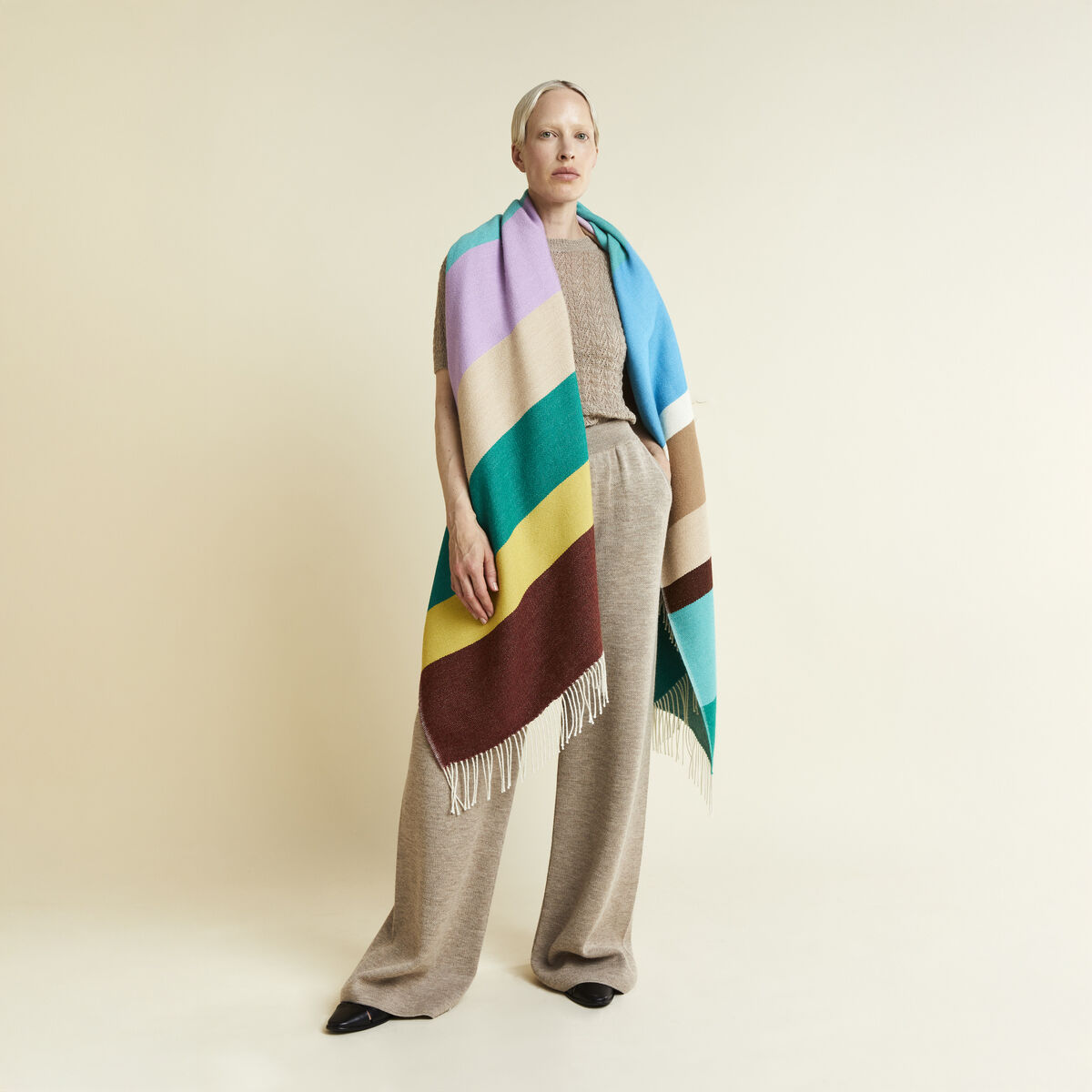 Das Bild zeigt eine Frau mit dem Cape »Mainau« von parati aus 100% Alpaka in Überlänge. Das Cape ist gestreift in Beige, Braun, Türkis, Gelb, Lila und Grün, mit Fransen an den Enden. Die Frau trägt beige Kleidung vor neutralem Hintergrund in elegantem Stil.