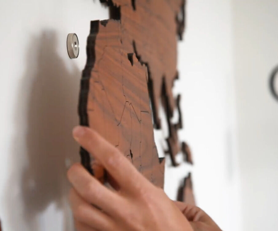 Nahaufnahme einer Holz-Weltkarte aus Nussbaumholz, die an eine weiße Wand angebracht wird. Eine Hand fixiert einen Teil der Karte an der Wand.
