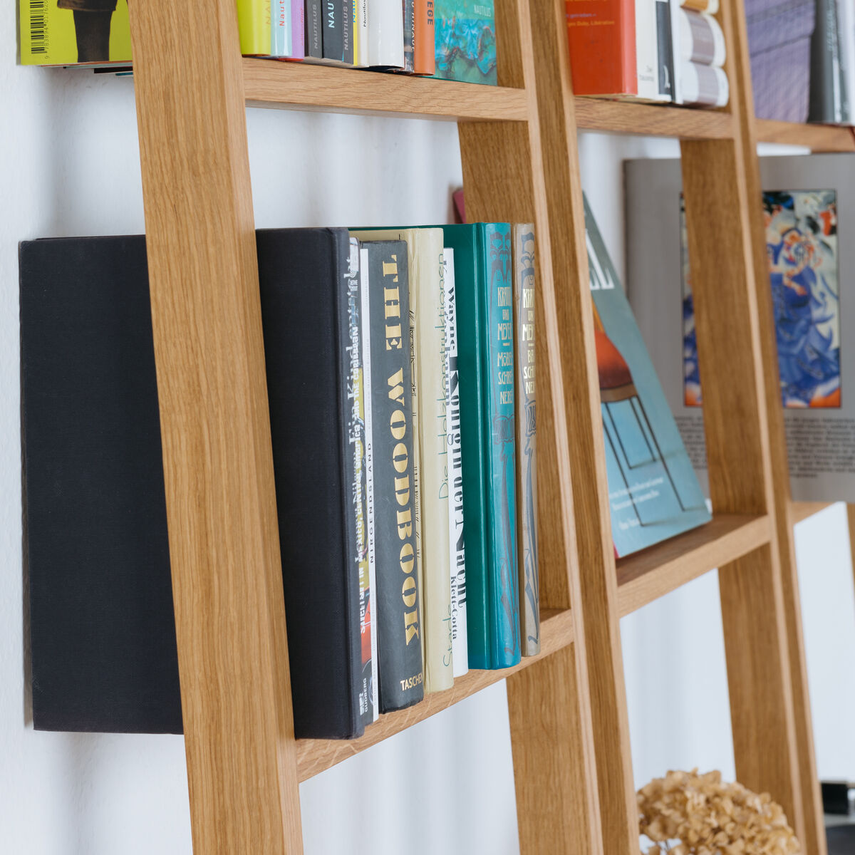 Ein Leiterregal namens »brecht« aus Holz, das an einer Wand aufgestellt ist. Das Regal ist mit mehreren Büchern und Dekorationselementen bestückt. Das Bild zeigt einen Ausschnitt des Regals im Kontext eines modern eingerichteten Raumes mit weiß gestrichener Wand.