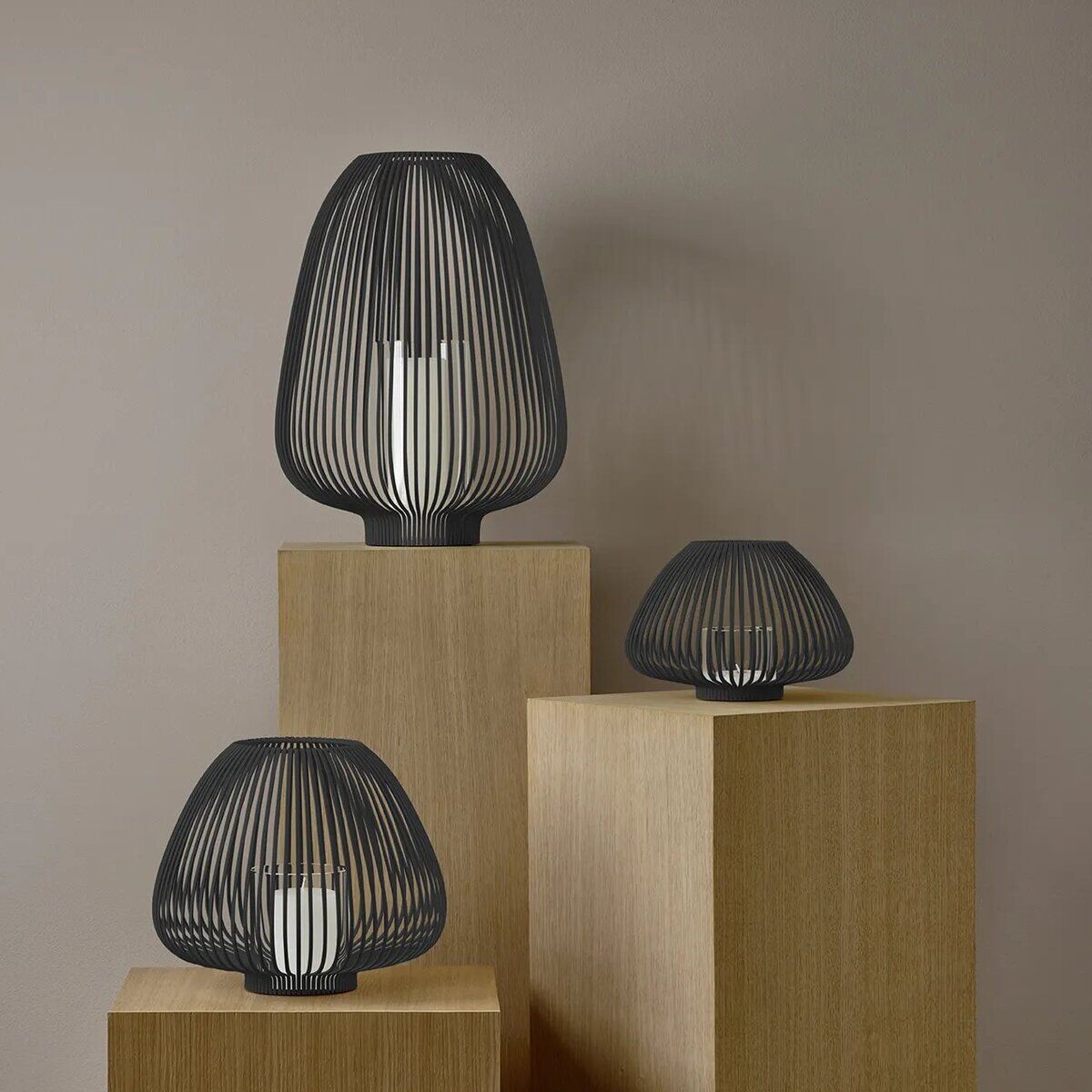 Drei moderne Laternen aus schwarzem Metall mit Gitterdesign stehen auf unterschiedlich hohen, rechteckigen Holzsockeln vor einer beigefarbenen Wand. Jede Laterne enthält eine weiße Kerze und strahlt einen minimalistischen, stilvollen Charakter aus, passend für Wohnbereich, Terrasse oder als dekoratives Element zu besonderen Anlässen.