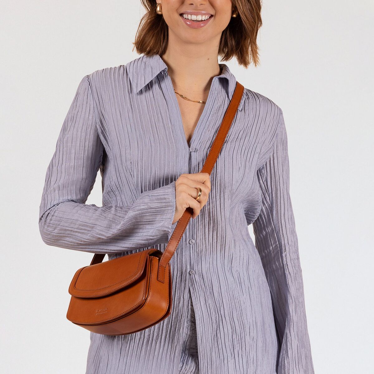 Eine Frau trägt eine kleine, braune Umhängetasche namens »Kendalini« über der Schulter. Sie trägt ein hellblaues, langärmliges Kleid mit feinen Falten. Die Tasche ist schlicht und elegant und eignet sich gut für den täglichen Gebrauch oder als modisches Accessoire.