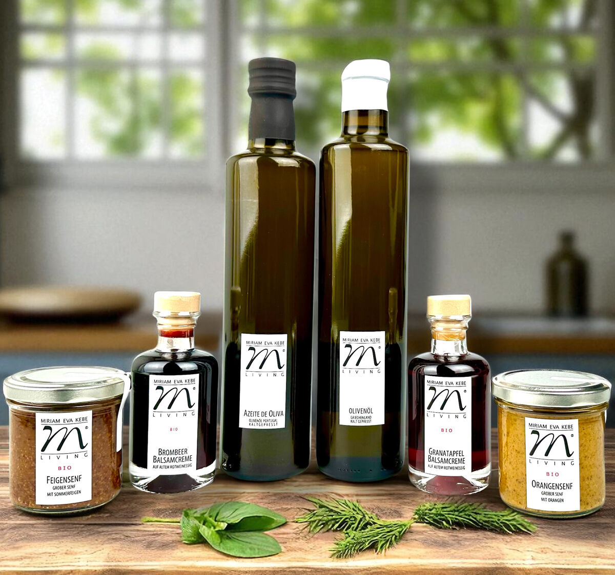 Mehrteiliges Gourmet-Set bestehend aus zwei braunen Flaschen Olivenöl und Essig, drei kleinen Gläsern mit Aufstrichen und zwei Kräuterzweigen, arrangiert auf einem Holztisch vor einem Fenster mit Gartenblick. Ideal als Geschenkset für Feinschmecker und zum Kochen oder Genießen besonderer Speisen.