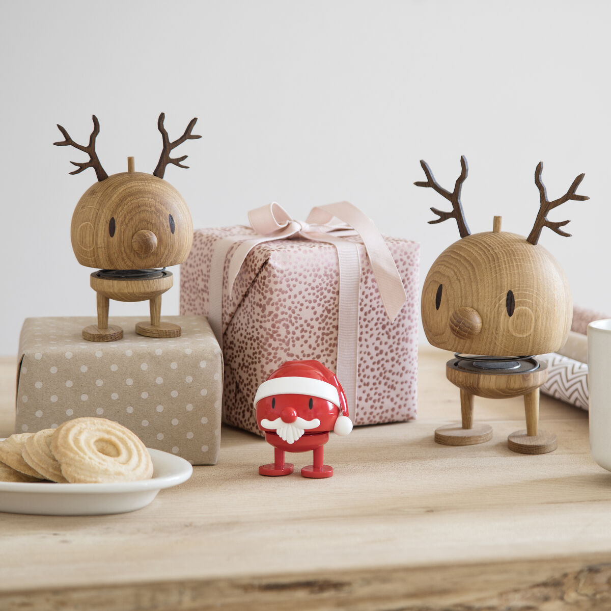Zwei hölzerne Rentierfiguren der Marke Hoptimist stehen auf einem hellen Holztisch, begleitet von liebevoll verpackten Geschenken, einer weißen Tasse, Gebäck und einer kleinen rot-weißen Weihnachtsfigur vor neutralem Hintergrund. Die Szene vermittelt eine festliche, skandinavische Weihnachtsatmosphäre.