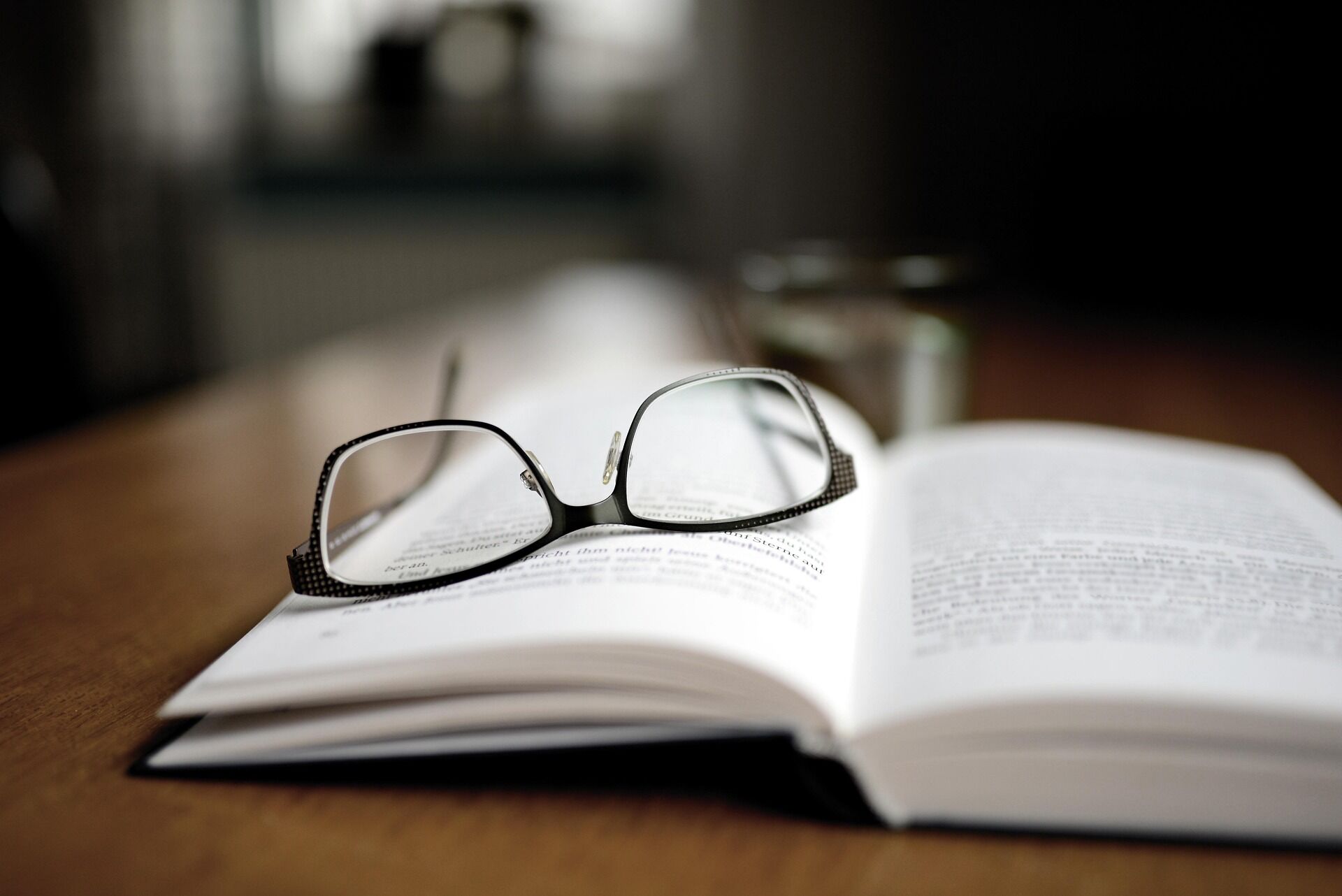 Link zur Kategorie Bücher, um die Welt zu verstehen. Nahaufnahme eines aufgeschlagenen Buchs auf einem Holztisch, mit einer schwarzen Lesebrille auf den Seiten. Im Hintergrund steht unscharf ein Glas Wasser.