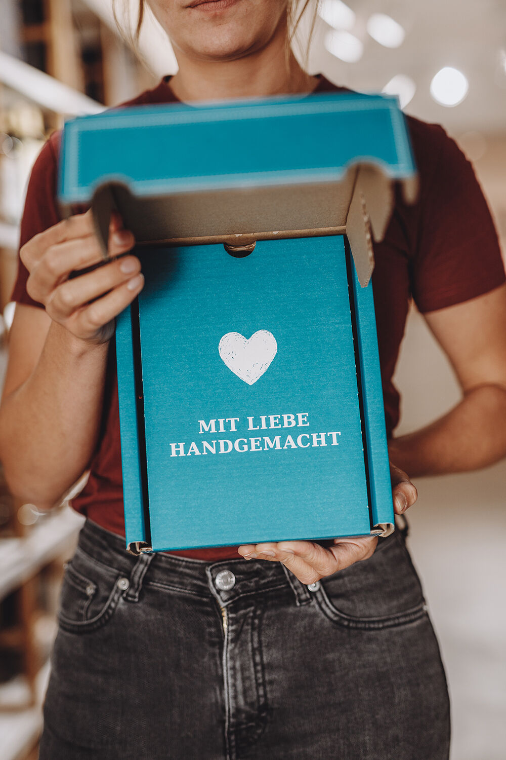 Person hält geöffneten blauen Geschenkkarton mit weißem Herz und Schrift 'MIT LIEBE HANDGEMACHT'