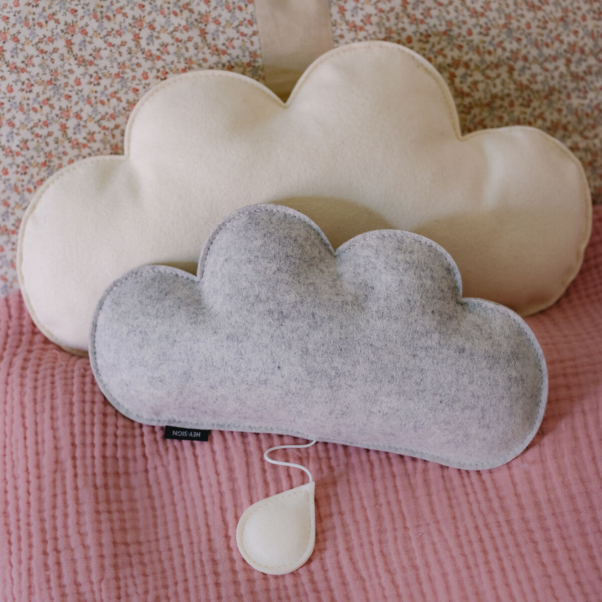 Spieluhr in Form einer Wolke mit grau-weißem Plüschstoff auf rosafarbener, gesteppter Decke. Hintergrund mit beigefarbenem, gemustertem Stoff.