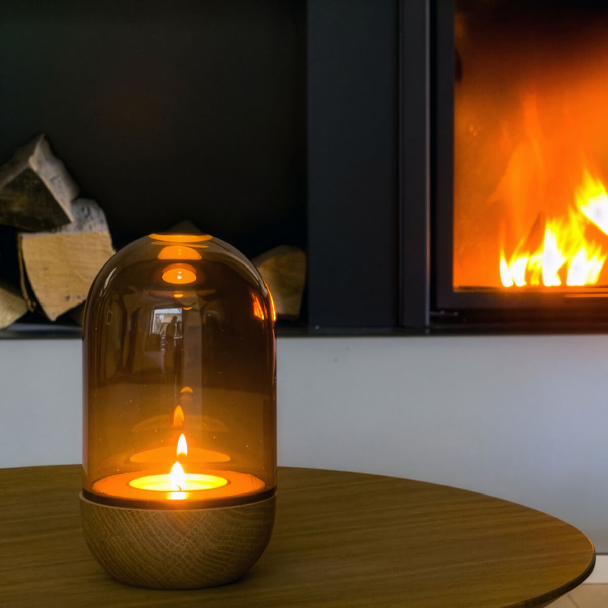 Windlicht »GLOW« der Marke finbeaux aus Rauchglas auf rundem Holztisch, brennende Kerze im Inneren. Im Hintergrund ein moderner Kamin mit sichtbaren Flammen und gestapeltem Holz. Stilvolle, warme Atmosphäre im Wohnzimmer-Setting.