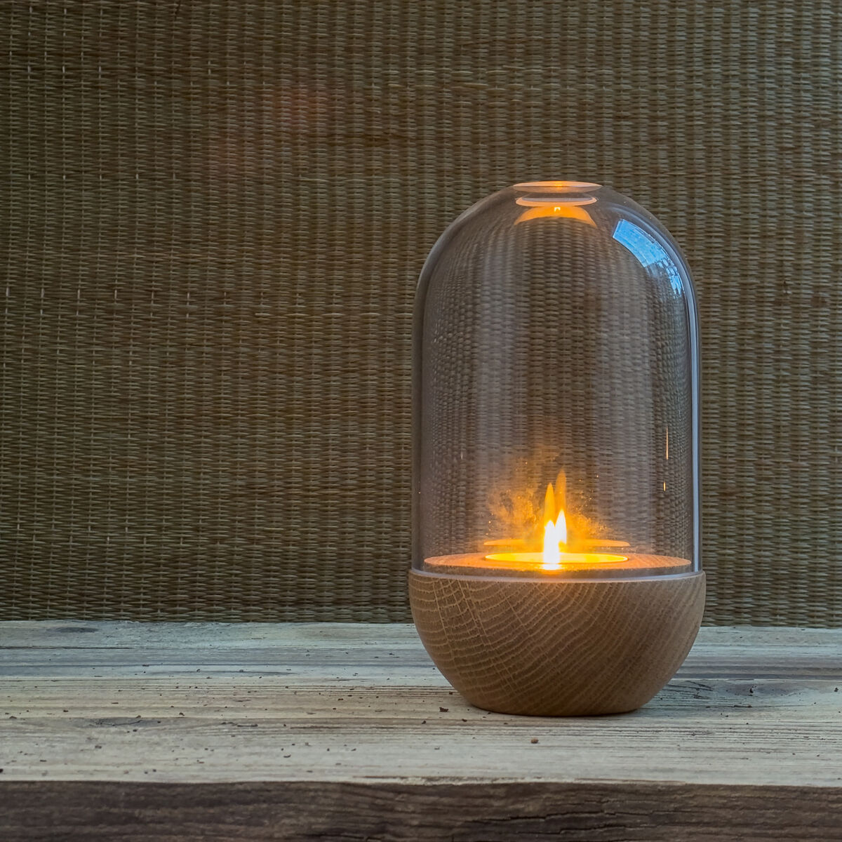 Windlicht »GLOW« von finbeaux, bestehend aus einer massiven Eichenbasis und einer mundgeblasenen Klarglasglocke. Im Inneren brennt eine Flamme, die ein warmes Licht wirft. Das Windlicht steht auf einer Holzfläche vor einem strukturierten braunen Hintergrund.